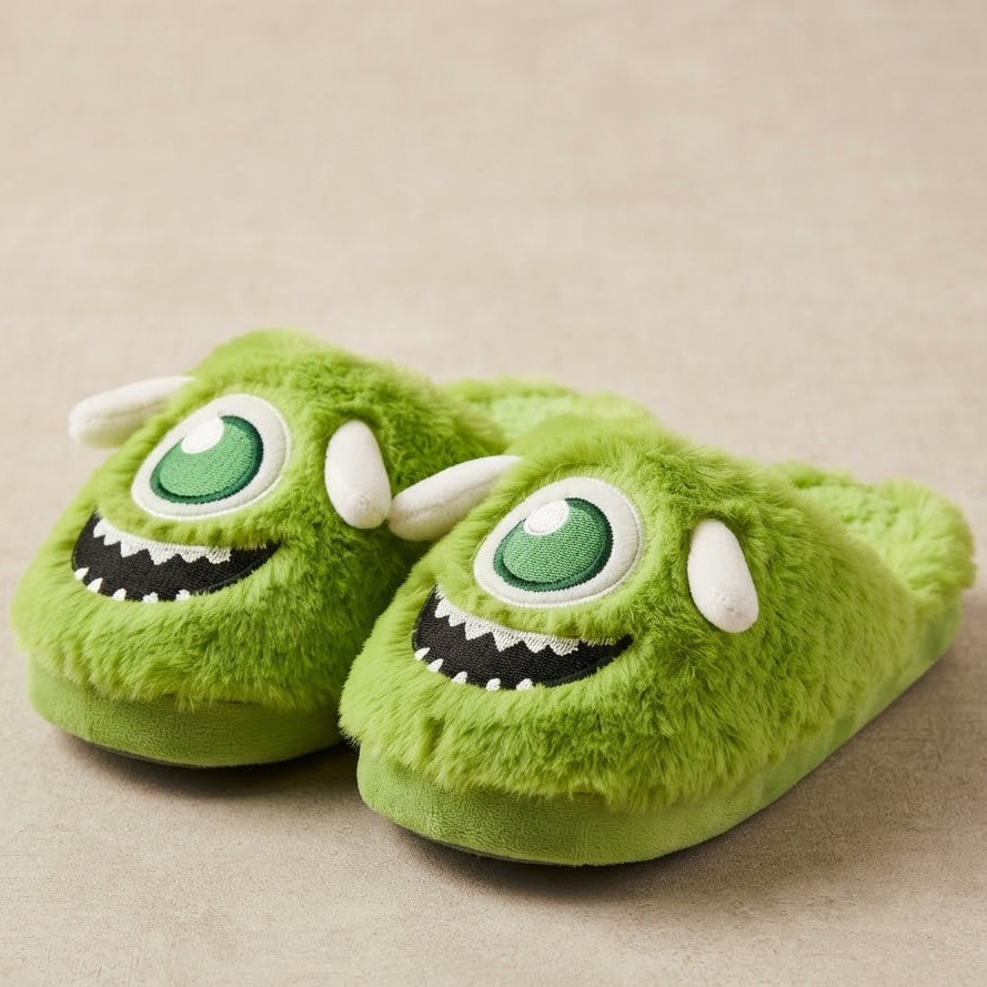 A pair of plush green monster-themed slippers with large embroidered eyes, white horns, and a toothy grin, photographed on a neutral beige background.