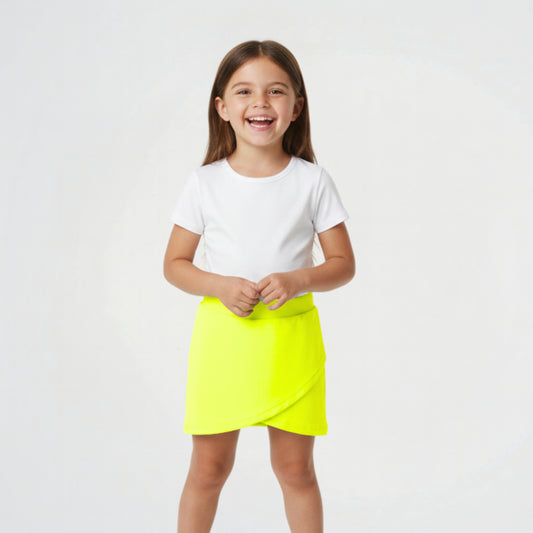 A young girl smiling and wearing a white short-sleeve t-shirt and a bright neon yellow wrap-style mini skirt, standing against a plain light gray studio background.