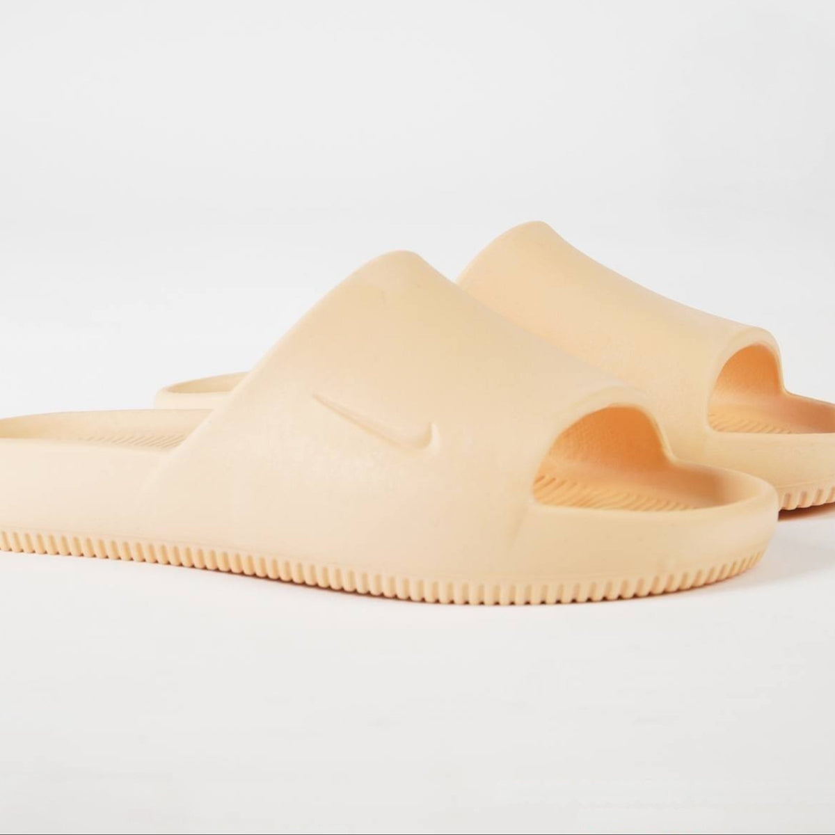 A pair of chunky, open-toe slide sandals in a solid pale peach-beige color, featuring a molded Nike Swoosh on the strap and a textured ridged sole, set against a clean white background.