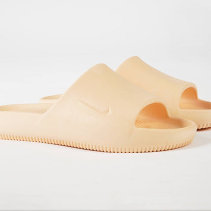 A pair of chunky, open-toe slide sandals in a solid pale peach-beige color, featuring a molded Nike Swoosh on the strap and a textured ridged sole, set against a clean white background.