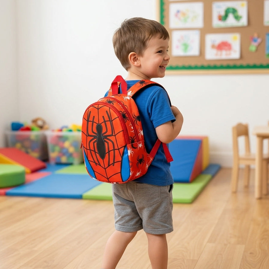 A young boy wearing a red and blue Spider-Man themed backpack, blue t-shirt, and gray shorts walks in a brightly lit children's playroom. The backpack features a large black spider graphic on a red background with blue side panels.
