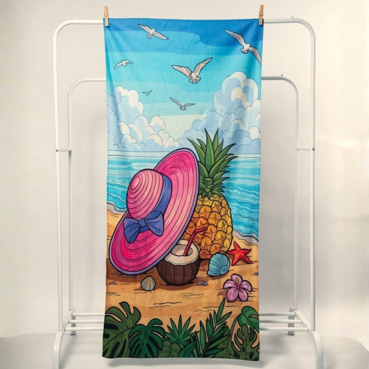 A beach towel featuring a colorful tropical illustration of a pink sun hat, pineapple, coconut drink, starfish, ocean, and seagulls, hanging from a white metal stand.