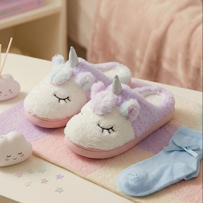 A pair of plush unicorn slippers in pastel lavender and cream with pink soles, displayed on a pastel blanket beside folded light blue socks and small cloud-shaped accessories on a tabletop.